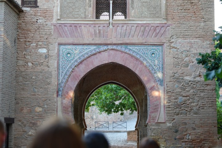 People standing before a historic stone archway with intricate carvings and a tree visible through it.