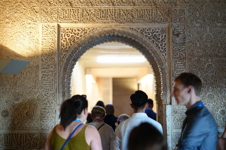 People walking through ornate stone archway with intricate carvings in dim lighting.