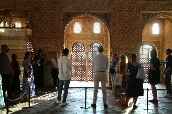 Group of people inside an ornate, sunlit, historical building with arched windows and patterned walls.